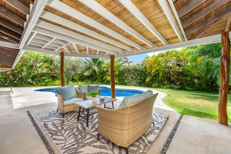 Covered wood-beam lanai with outdoor seating next to a pool and tropical yard